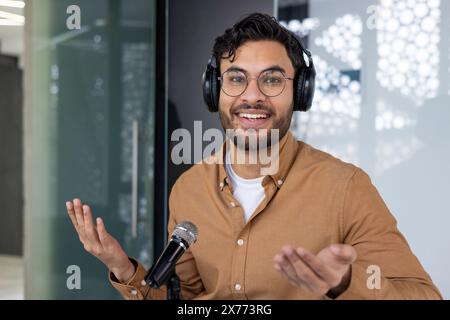 Un jeune homme joyeux portant des écouteurs et des lunettes, animant un podcast dans un studio moderne. Il semble énergique et engagé avec son public. Banque D'Images