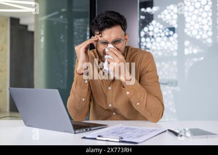 Homme aux prises avec des symptômes du rhume tout en essayant de travailler sur son ordinateur portable au bureau. Concept de maladie, problèmes de santé et environnement de travail. Banque D'Images