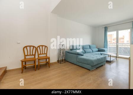 Salon avec un grand canapé recouvert de tissu bleu, quelques chaises en bois verni et un balcon avec des rideaux assortis Banque D'Images