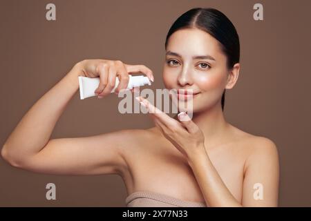 Belle femme avec bouteille de crème hydratante. Vidéo de femme avec la peau parfaite sur fond beige. Concept de beauté et de soins de la peau Banque D'Images