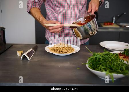 Homme en gros plan versant de la sauce tomate sur une assiette avec des spaghettis frais, debout à la table de la cuisine avec des ingrédients biologiques frais. Cuisson des pâtes italiennes selon Banque D'Images