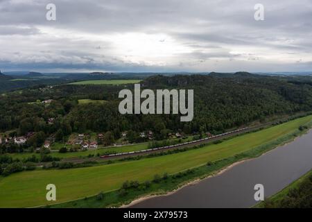 Un train descend une voie ferrée près d'une rivière. Le train est long et a une couleur rouge. Le ciel est nuageux, et l'atmosphère est calme et poac Banque D'Images