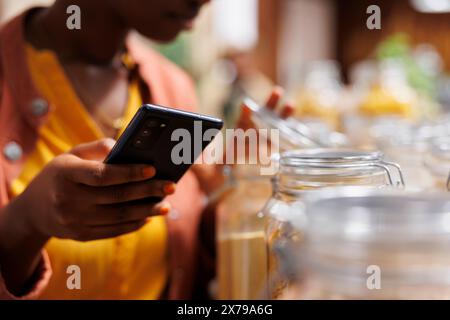 Dans cette image en gros plan, une femme noire tient un appareil mobile tout en vérifiant les articles dans les récipients en verre au magasin bio-alimentaire. Focus sélectif sur smartphone saisi par une afro-américaine. Banque D'Images