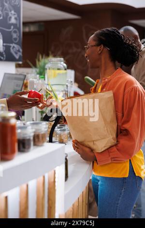 Image d'une femme noire avec un sac d'épicerie remettant du poivron rouge au vendeur local afro-américain. Une cliente souriante offrant ses produits frais sélectionnés à la caissière, shopping durable. Banque D'Images