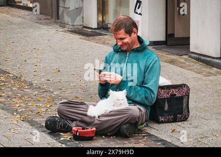 Un homme souriant est assis les jambes croisées dans une rue luxembourgeoise avec un chat blanc sur ses genoux, regardant son téléphone, à côté d'un porte-animaux et d'un bol de don. Banque D'Images