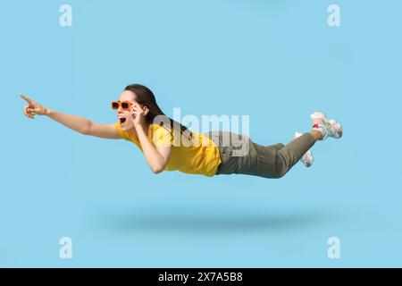 Surprise jeune femme dans des lunettes de soleil volant et pointant vers quelque chose sur fond bleu Banque D'Images