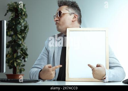 Homme confiant avec des cheveux gris piquants et des lunettes de soleil pointe à un tableau blanc vierge. Son costume gris clair et sa chemise noire, combinés avec son look souillé, sugg Banque D'Images