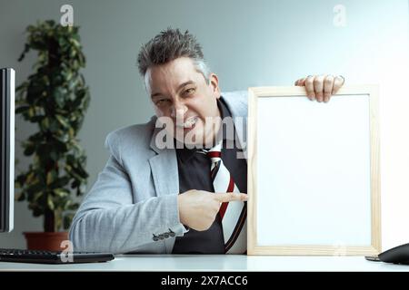 Homme d'âge moyen intense avec des pointes de cheveux grises pointues avec force sur un tableau blanc vierge. Son expression agressive contraste avec son costume gris clair et Banque D'Images