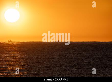 Soleil d'aube jaune émergeant de l'horizon rétro de la mer Méditerranée illuminant deux bateaux de pêche en silhouette allant pêcher des crevettes, sous un o Banque D'Images