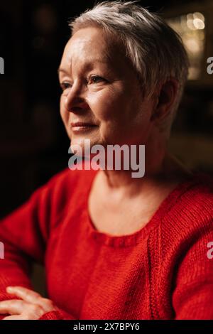 Gros plan vertical de la femme âgée aux cheveux gris positive dans un pull rouge décontracté assis à table dans une pièce sombre par la lumière de la bougie allumée. Banque D'Images