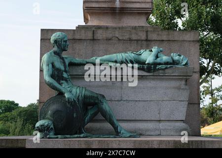 Statue en bronze représentant un homme assis tenant un bouclier et une figure couchée sur un piédestal de pierre dans un cadre extérieur. Banque D'Images