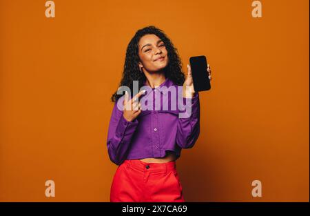 Jeune femme en vêtements décontractés se tient devant un fond orange. Elle montre un smartphone avec un écran tactile dans une main. Banque D'Images