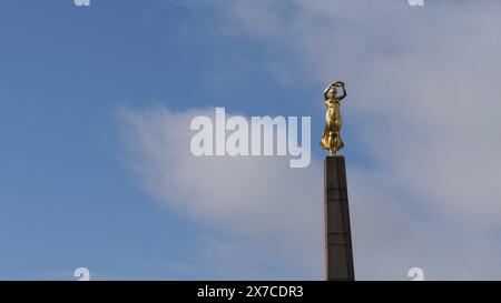 Statue dorée d'une femme tenant une couronne au sommet d'une haute colonne contre un ciel bleu avec des nuages. Banque D'Images