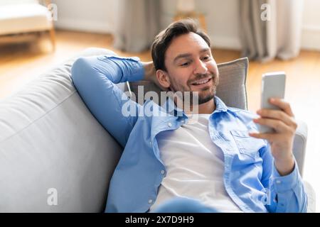 Homme positif assis sur le canapé regardant téléphone Banque D'Images