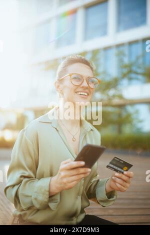 employé féminin moderne souriant près du centre d'affaires dans une blouse verte et des lunettes avec carte de crédit à l'aide d'un smartphone. Banque D'Images