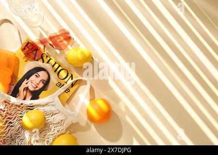 Sac à ficelle avec des fruits frais, magazine de mode et accessoires de plage sur fond beige, pose plate. Espace pour le texte Banque D'Images