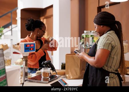 Après avoir payé pour des produits bio-alimentaires qu'elle a sélectionnés dans un magasin respectueux de l'environnement, une femme noire remet sa carte de crédit dans sa poche. Au comptoir de caisse, le commerçant et le client effectuent une transaction sans espèces. Banque D'Images