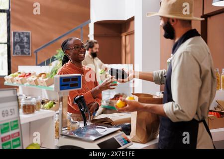 Cliente afro-américaine souriante qui remet des aubergines cultivées localement à un caissier du moyen-Orient pour peser. Femme noire aidée par un vendeur masculin à la caisse. Banque D'Images