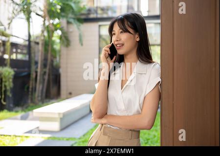 Une femme d'affaires asiatique millénaire attrayante et positive est appuyée contre le mur du bâtiment à l'extérieur et parle au téléphone avec quelqu'un. Banque D'Images