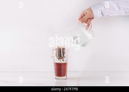 Tasse de café fraîchement infusé, rapide et instantanée moderne, sachet infusé café frais espresso dans une tasse en verre. Mains de femme versant de l'eau dans le sac filtre à café Banque D'Images