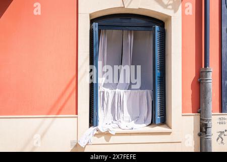 Fenêtre avec volets bleus sur un bâtiment avec façade blanche et rose. Des rideaux blancs pendent d'une fenêtre dans un vieux bâtiment. Banque D'Images