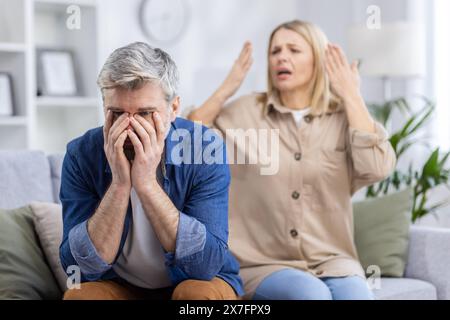 Homme frustré couvrant son visage avec ses mains tandis que son partenaire fait des gestes en arrière-plan lors d'une dispute animée dans leur salon. Banque D'Images