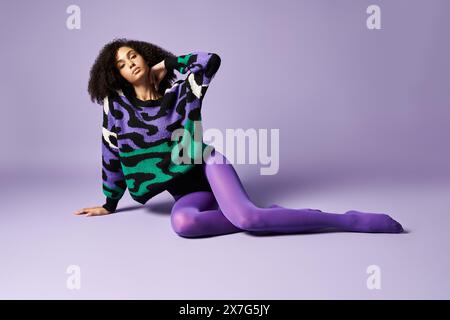 Une jeune femme portant des collants violets et un pull assis sur le sol dans un studio avec un fond violet. Banque D'Images
