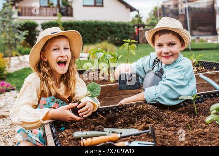 Fille et garçon prenant soin de petites plantes de légumes dans un lit surélevé, tenant une petite pelle. Enfance en plein air dans le jardin. Banque D'Images