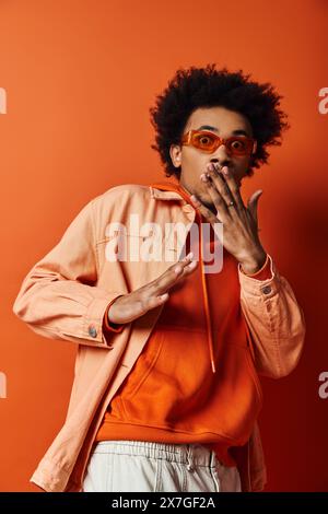 Un jeune homme afro-américain branché dans une chemise orange et des lunettes de soleil tient ses mains jusqu'à sa bouche dans un geste d'émotion. Banque D'Images