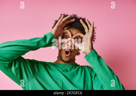 Un jeune homme afro-américain en tenue décontractée sur un fond rose, faisant un visage ludique et expressif en utilisant ses mains. Banque D'Images