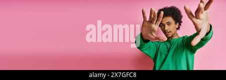 Joyeux homme afro-américain bouclé en chemise verte décontractée avec les mains levées, exprimant la positivité sur un fond rose. Banque D'Images