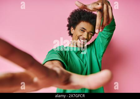 Un homme afro-américain joyeux, cheveux bouclés dans une chemise verte, danse énergiquement sur un fond rose éclatant. Banque D'Images