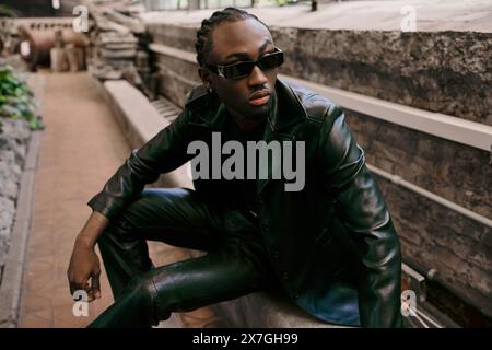 Bel homme afro-américain en veste en cuir et lunettes de soleil est assis en toute confiance sur le rebord dans le jardin vert vif. Banque D'Images