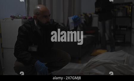 Un homme chauve examine des preuves sur une scène de crime à l'intérieur avec un corps couvert alors qu'une femme enquête en arrière-plan. Banque D'Images