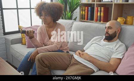 Couple interracial se prélassant sur un canapé, avec la femme tenant une télécommande dans un salon confortable Banque D'Images Couple interracial se prélassant sur un canapé, avec la femme tenant une télécommande dans un salon confortable Banque D'Images