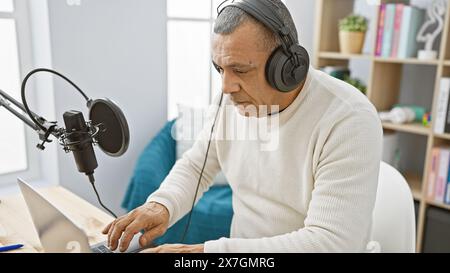Homme hispanique mature avec des écouteurs utilisant un ordinateur portable dans l'intérieur du studio de radio. Banque D'Images