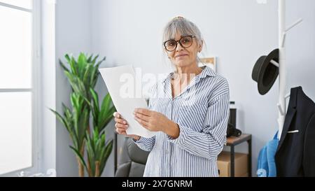 Femme mature avec des lunettes dans la chemise rayée tient le papier dans le bureau moderne, représentant la vie quotidienne des affaires. Banque D'Images