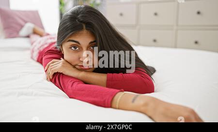 Une jeune femme indienne sereine aux cheveux longs se détend dans une chambre lumineuse allongée sur un lit blanc Banque D'Images