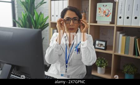 Une femme hispanique d'âge moyen réfléchie portant une blouse de laboratoire blanche ajuste ses lunettes dans un lieu de travail de clinique, reflétant la concentration et le professionnalisme. Banque D'Images