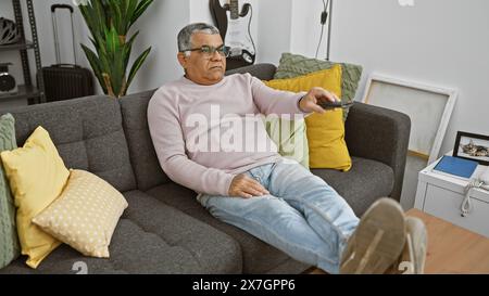 Homme mûr se relaxant avec télécommande sur le canapé dans l'intérieur confortable du salon, concept de loisirs. Banque D'Images