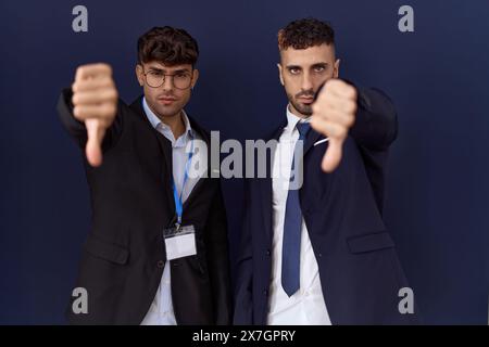 Deux hommes d'affaires hispaniques portant des vêtements d'affaires l'air malheureux et en colère montrant le rejet et le négatif avec le geste pouce vers le bas. mauvaise expression. Banque D'Images