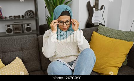 Une femme mature dans un hijab lit intentionnellement un livre tout en ajustant ses lunettes, assise sur un canapé à l'intérieur. Banque D'Images