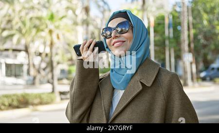 Une élégante femme d'âge moyen portant des lunettes de soleil et un hijab écoute un message vocal sur son smartphone à l'extérieur. Banque D'Images