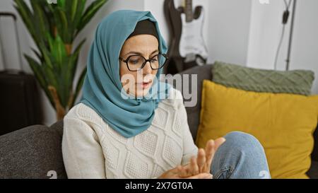 Une femme hispanique mature portant des lunettes et un hijab ressent des douleurs aux mains dans un cadre confortable de salon. Banque D'Images