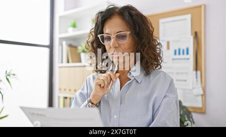 Pensive hispanique femme d'affaires examine des documents dans un cadre de bureau moderne, incarnant l'expertise et le professionnalisme. Banque D'Images