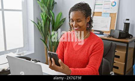 Femme afro-américaine en tenue professionnelle utilise smartphone dans un cadre de bureau moderne, transmettant la connectivité et la productivité. Banque D'Images