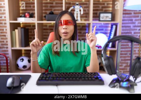 Femme chinoise d'âge moyen portant des lunettes de réalité virtuelle étonnée et surprise regardant vers le haut et pointant avec les doigts et levant les bras. Banque D'Images