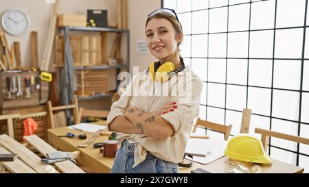 Femme confiante avec des tatouages se croise les bras dans un atelier de menuiserie ensoleillé portant des écouteurs. Banque D'Images