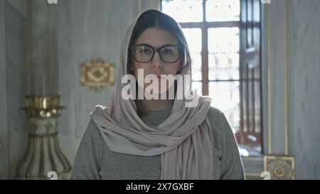 Une femme réfléchie portant des lunettes et un hijab pose à l'intérieur d'une mosquée, incarnant une atmosphère religieuse sereine. Banque D'Images
