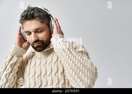 Un homme élégant avec une barbe porte un pull blanc, immergé dans la musique à travers des écouteurs, sur fond de studio gris. Banque D'Images